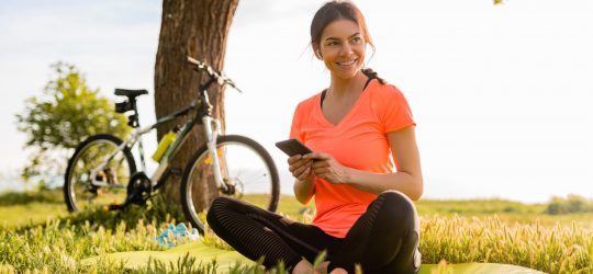smiling beautiful woman holding phone doing sports in morning in park nature on mat wearing pink shirt fitness outfit, happy healthy lifestyle, music in earphones, bicycle on background